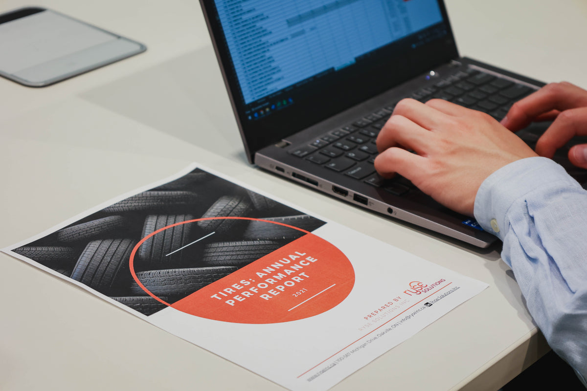 A person at a desk typing on a laptop with compliance data being reviewed on a screen. A printed report titled 'Tires Annual Performance Report' placed in the foreground on a white tabletop.