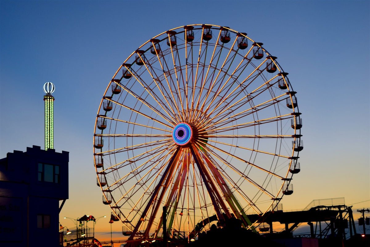 A large Ferris wheel at a fair or amusement park against a sunset sky, with a blue gradient background and silhouetted structures in the foreground. Farmer's markets, tournaments, festivals, converts and other types of events all require responsible and reliable waste removal solutions. 