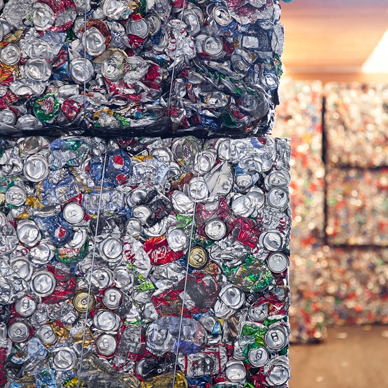 Large compressed bales of crushed aluminum and mixed recyclable cans stacked indoors, with a warm-toned warehouse or processing area in the background.