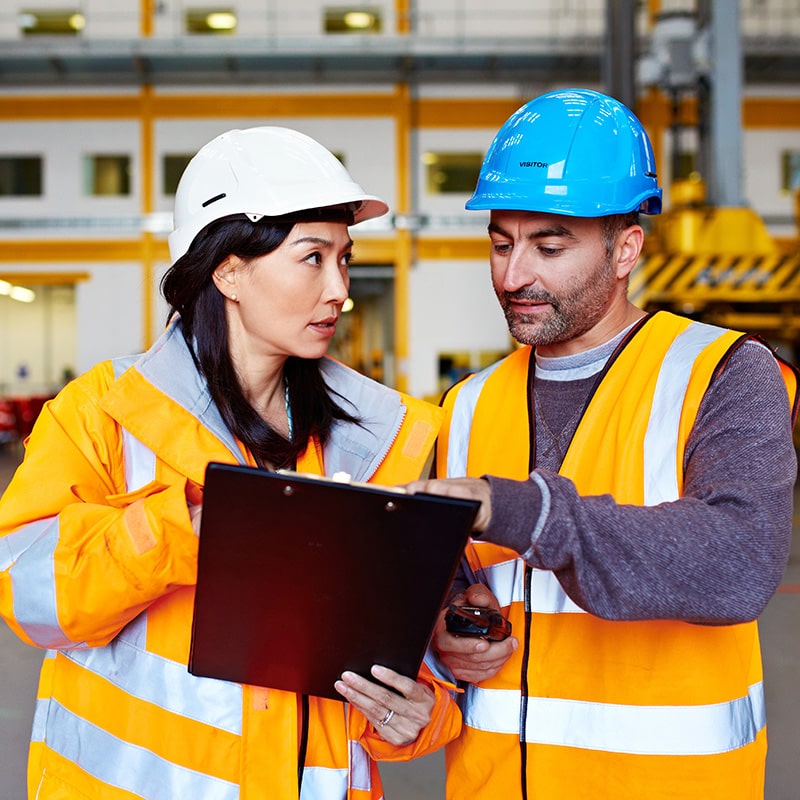 Two construction workers in high-visibility safety vests and hard hats standing in an industrial facility, with one holding a clipboard and the other pointing at it. Waste stream audits help customers better understand what's in their waste and discover new ways to divert and recycle more.